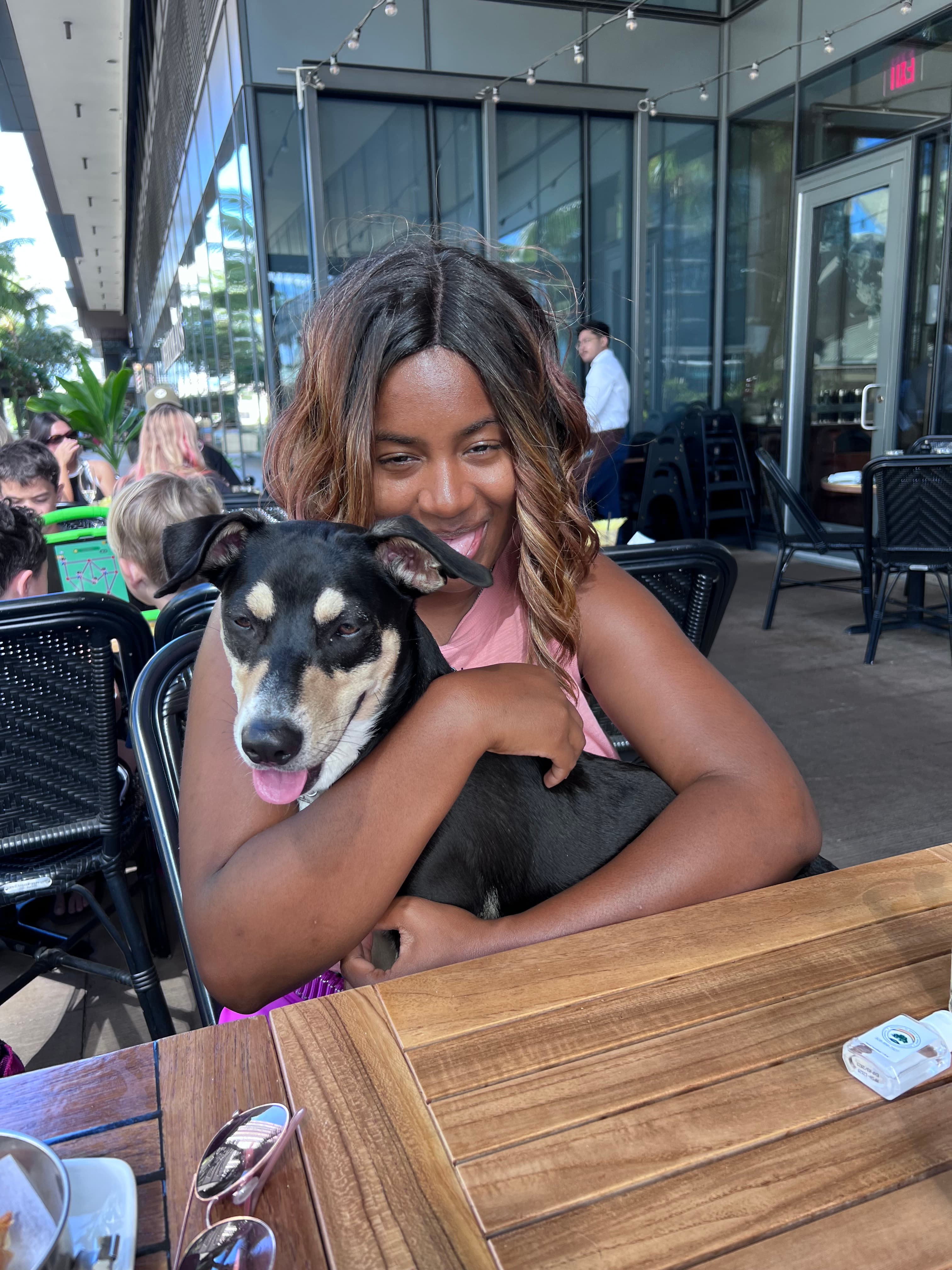 A smiling woman hugs a black and tan dog at an outdoor restaurant table.